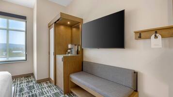 A modern hotel room with a large window, a sofa, a wooden cabinet, and a flat-screen TV mounted on the wall.