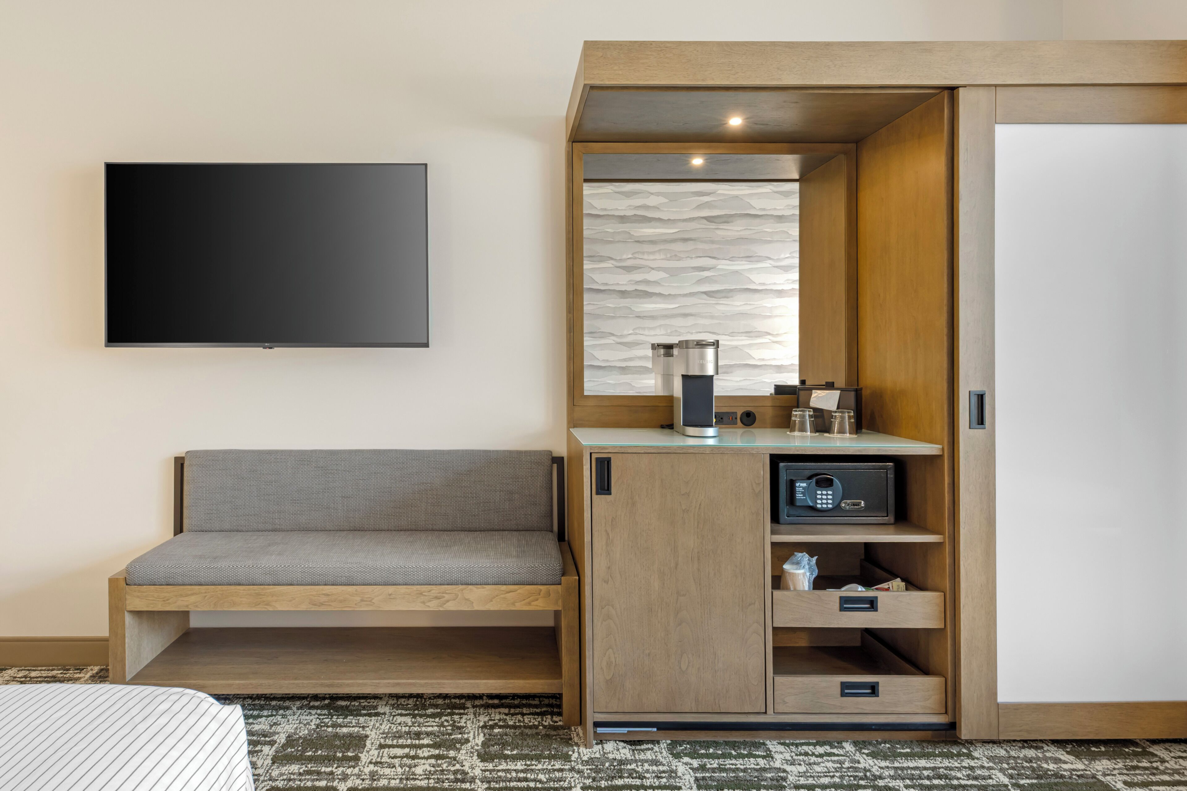 A hotel room with a flat-screen TV mounted on the wall, a wooden bench with storage drawers, a minibar, and a sliding door closet.