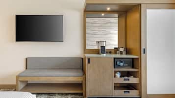 A hotel room with a flat-screen TV mounted on the wall, a wooden bench with storage drawers, a minibar, and a sliding door closet.