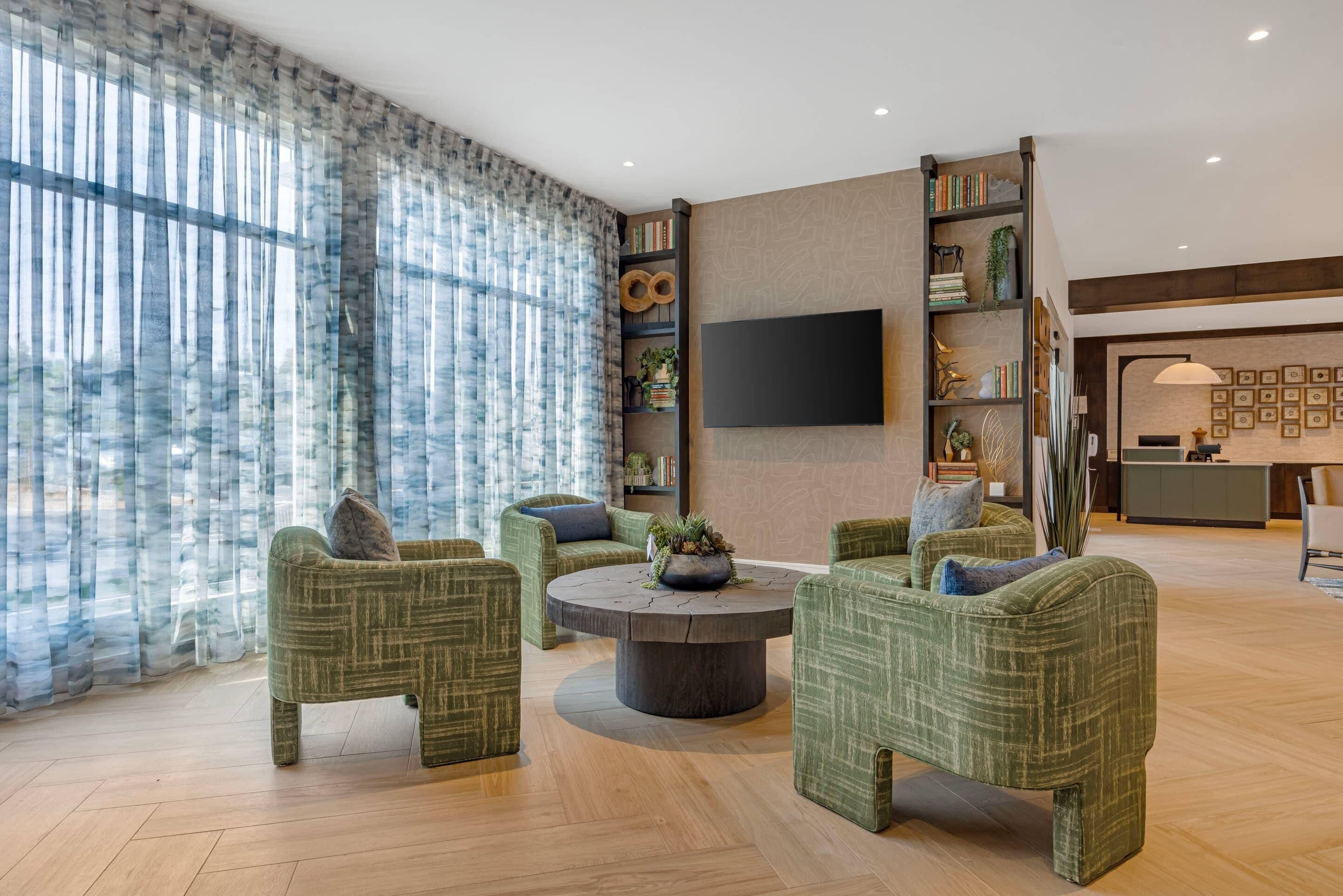 A modern living room with green upholstered chairs, a round coffee table, and a flat-screen TV mounted on the wall.