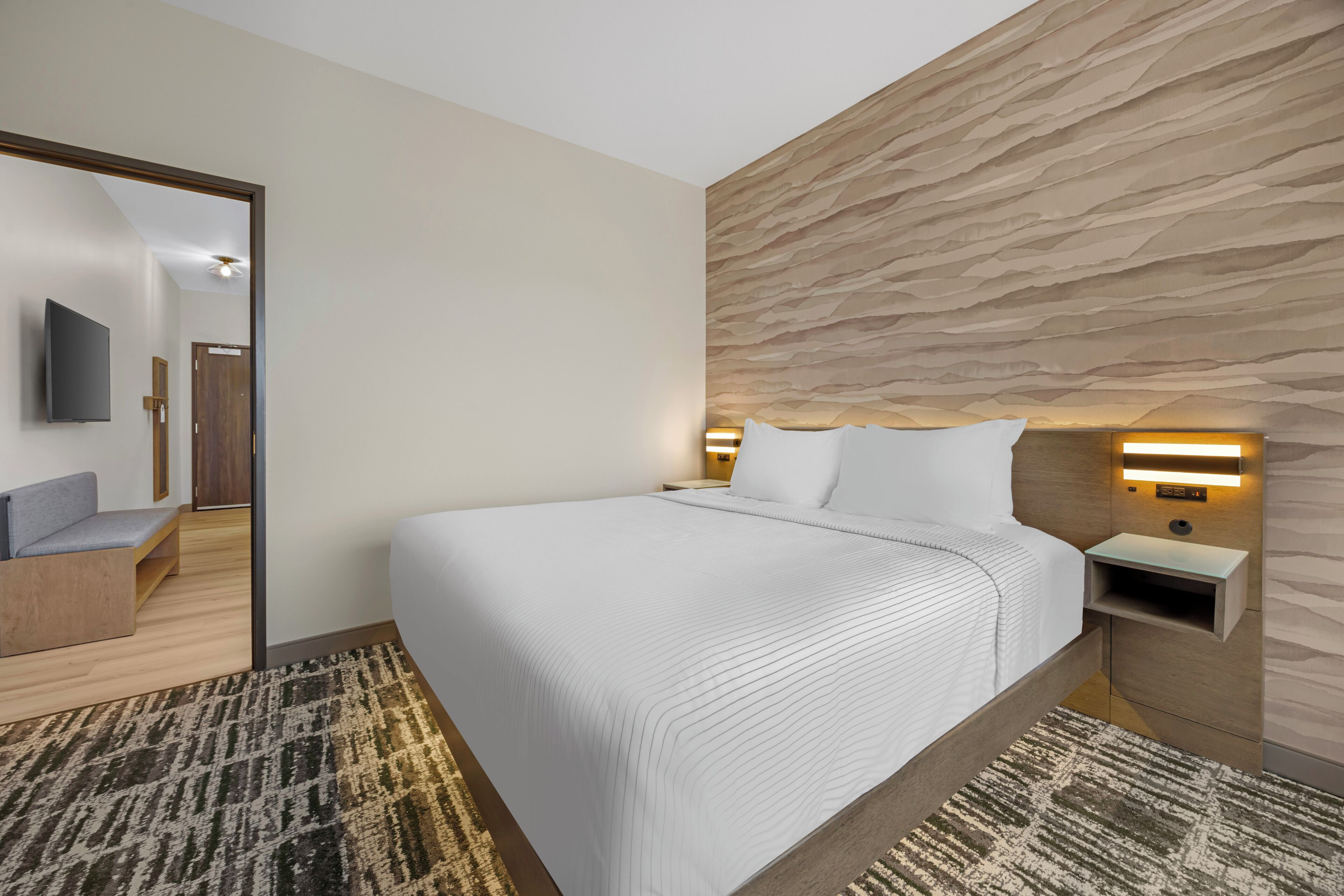 Una habitación de hotel moderna con una cama grande, un televisor de pantalla plana, cabecera de madera y un tapete estampado.