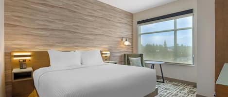 A hotel room with a large bed, a nightstand, a wall-mounted lamp, a chair, and a view of the outdoors through a window.