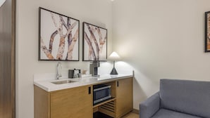 A compact kitchenette with a sink, stove, and microwave, a grey sofa, and two framed abstract paintings on the wall.