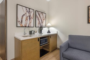 A compact kitchenette with a sink, stove, and microwave, a grey sofa, and two framed abstract paintings on the wall. - Cambria Hotel Bend - Mt. Bachelor (Bend)