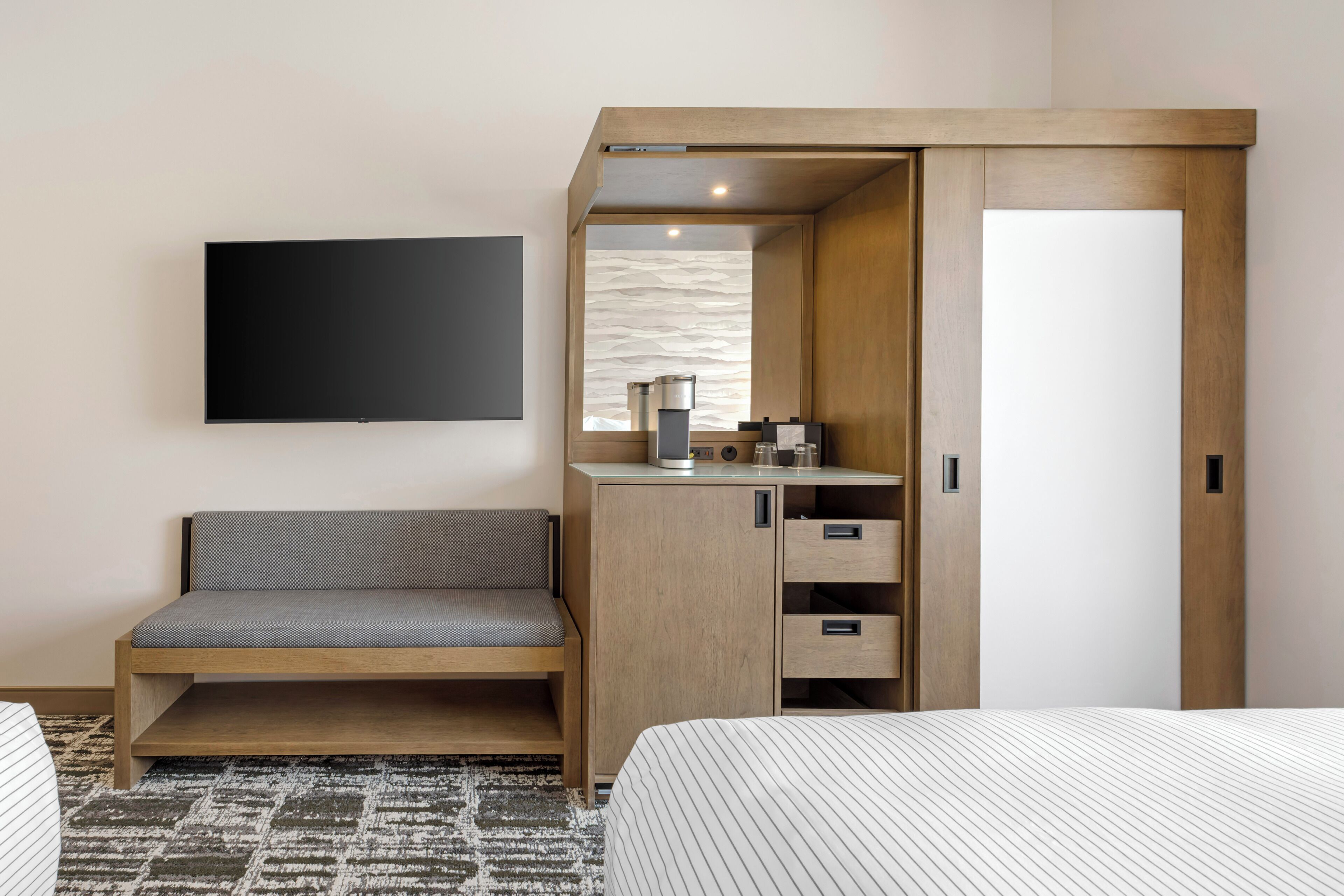 Habitación de hotel con cama, sofá, televisor montado en la pared, armario de madera y una mesita con tetera y tazas.