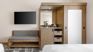 Habitación de hotel con cama, sofá, televisor montado en la pared, armario de madera y una mesita con tetera y tazas.