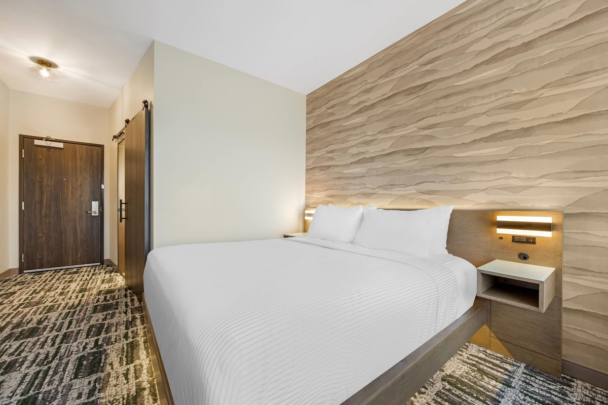 Habitación de hotel con una cama grande, cabecera de madera, mesita de noche y lámpara de pared.