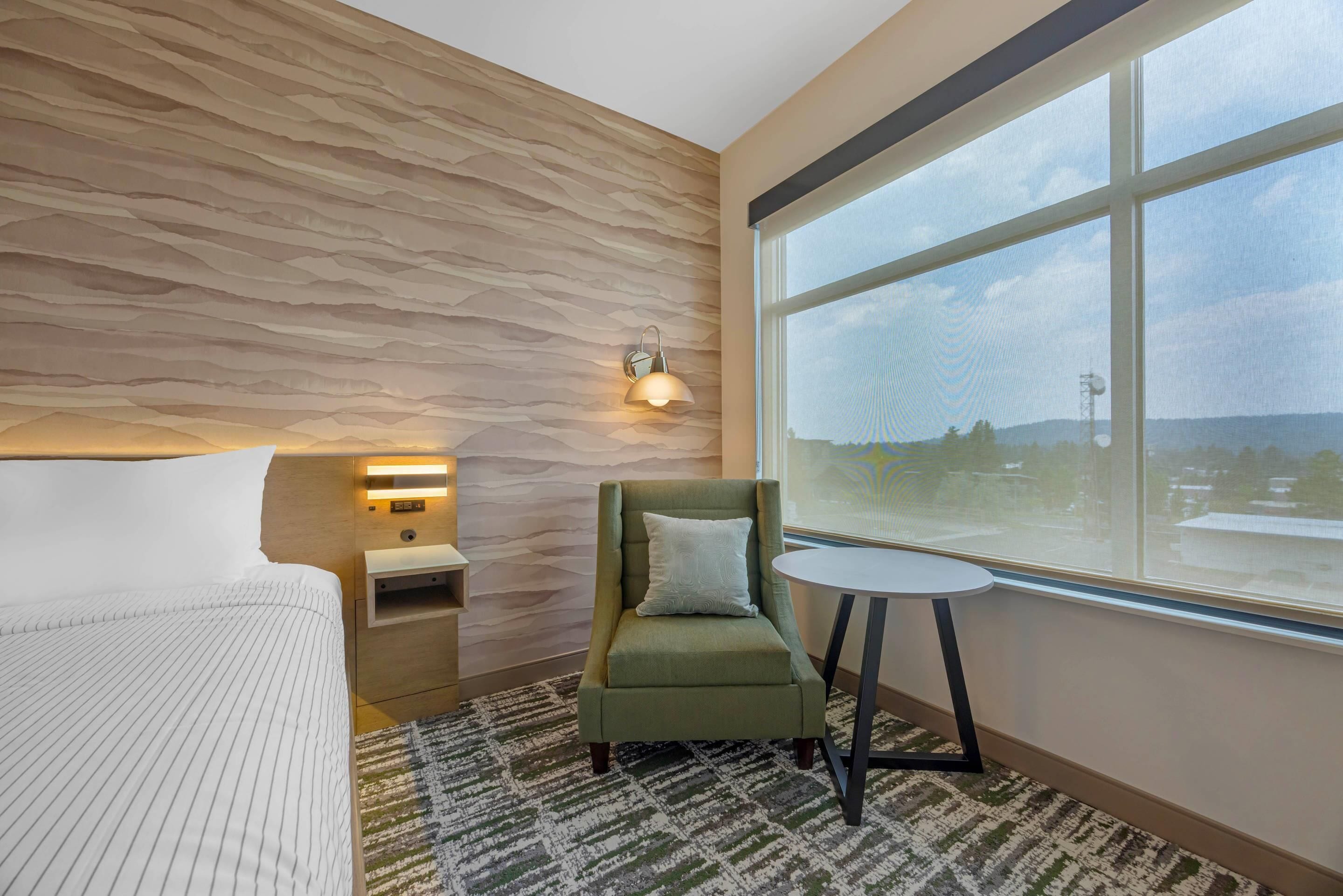 A hotel room with a bed, a green armchair, a small table, a nightstand, a wall-mounted lamp, and a large window with a view.