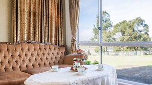 Ein Zimmer mit einem braunen Sofa mit Knöpfen, einem Tisch mit weißen Spitzen und einem Fenster mit Blick auf Bäume.