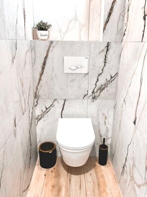 A bathroom with a white toilet, marble walls, a wooden floor, a black trash bin, and a small potted plant labeled 'Garden'. - Relax in Nature. Cozy Georgian resort with pool, stunning Views &Tranquility (Tskaltubo)