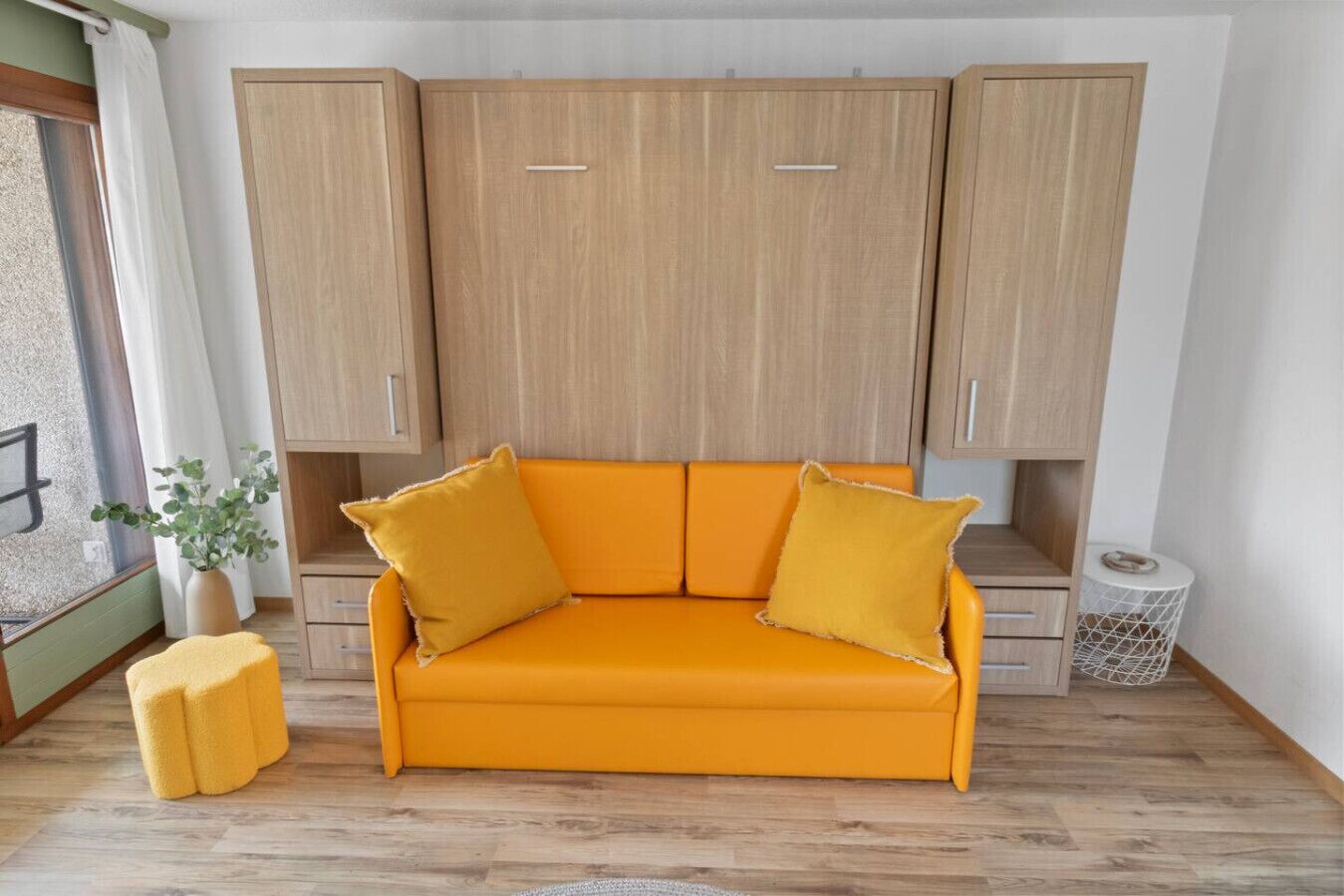 Una habitación con un armario de madera, un sofá amarillo con dos cojines, un pequeño banquillo amarillo y una planta en maceta.