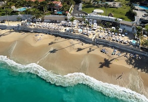 Vista aérea de um resort à beira-mar, com uma extensa praia de areia, área de piscina com guarda-sóis e algumas palmeiras.
