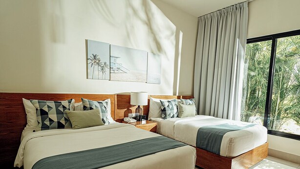 A hotel room with two beds, a nightstand, a lamp, and a large window with curtains. - Lifestyle Holiday Vacation Club (Puerto Plata)