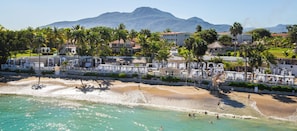 Un hotel frente a la playa con palmeras, una playa arenosa y una montaña al fondo.