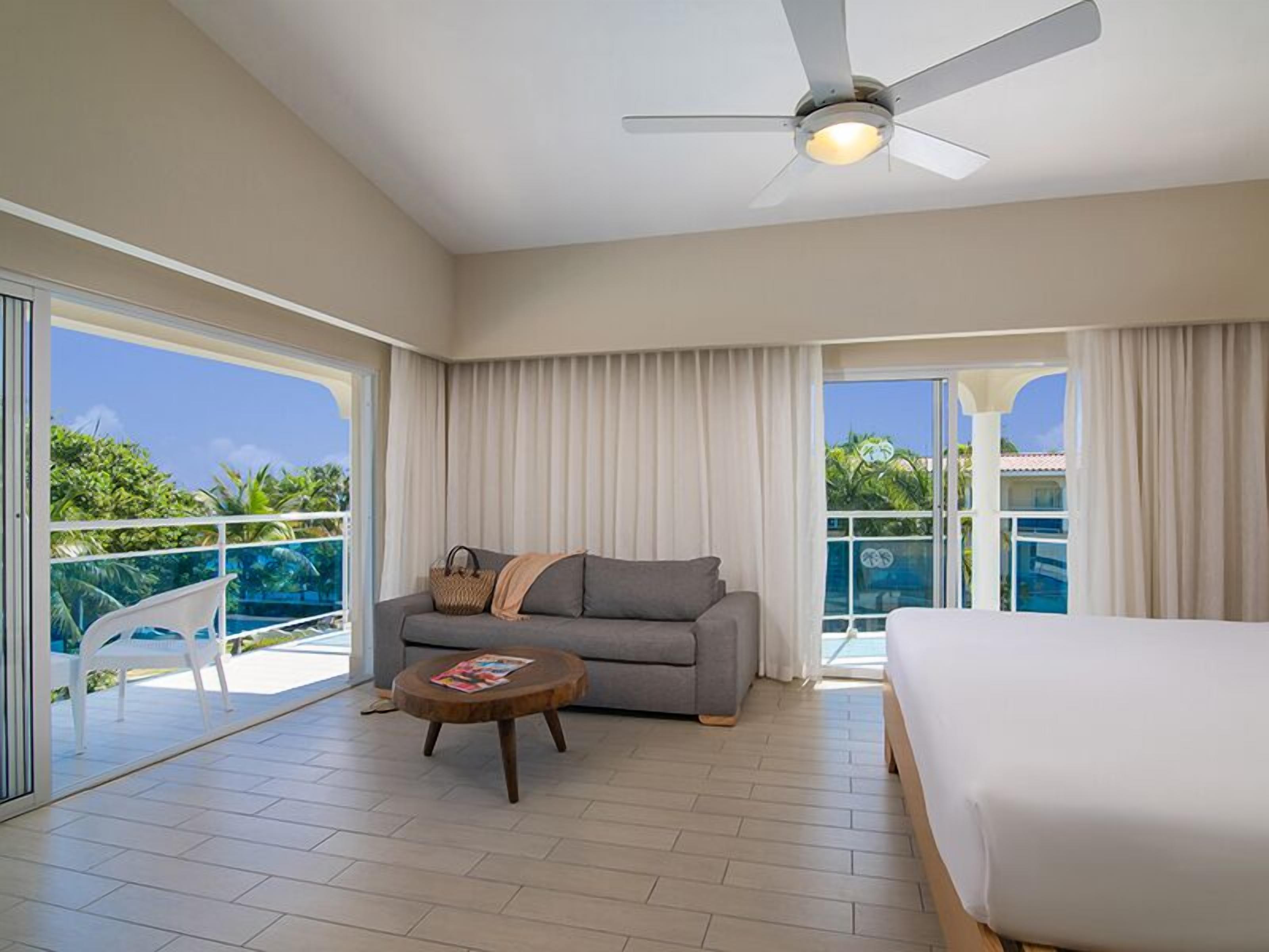A hotel room with a bed, a sofa, a small table, and a balcony with a view of the ocean.