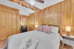 Ein Schlafzimmer mit Holzvertäfelung, einem Bett mit weißer Bettwäsche und Kissen in einem rosa Muster, einem Deckenventilator und einem Nachttisch mit Lampe.