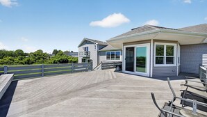 A covered outdoor deck with a wooden railing, a covered seating area, and a building with large windows and a door.