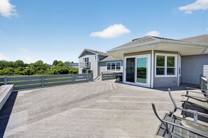 A covered outdoor deck with a wooden railing, a covered seating area, and a building with large windows and a door. - Chace Point by the Ocean (Little Compton)