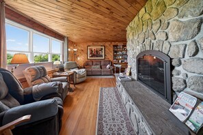A cozy living room with a stone fireplace, wooden ceiling, and large windows. - Chace Point by the Ocean (Little Compton)