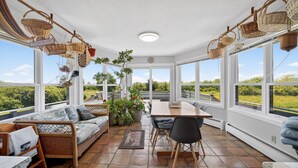 A sunlit room with wicker furniture, large windows, and a view of greenery.