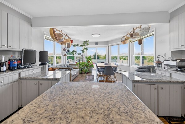 A kitchen with granite countertops, white cabinets, a dining area with a table and chairs, and large windows offering a view of the outdoors. - Chace Point by the Ocean (Little Compton)