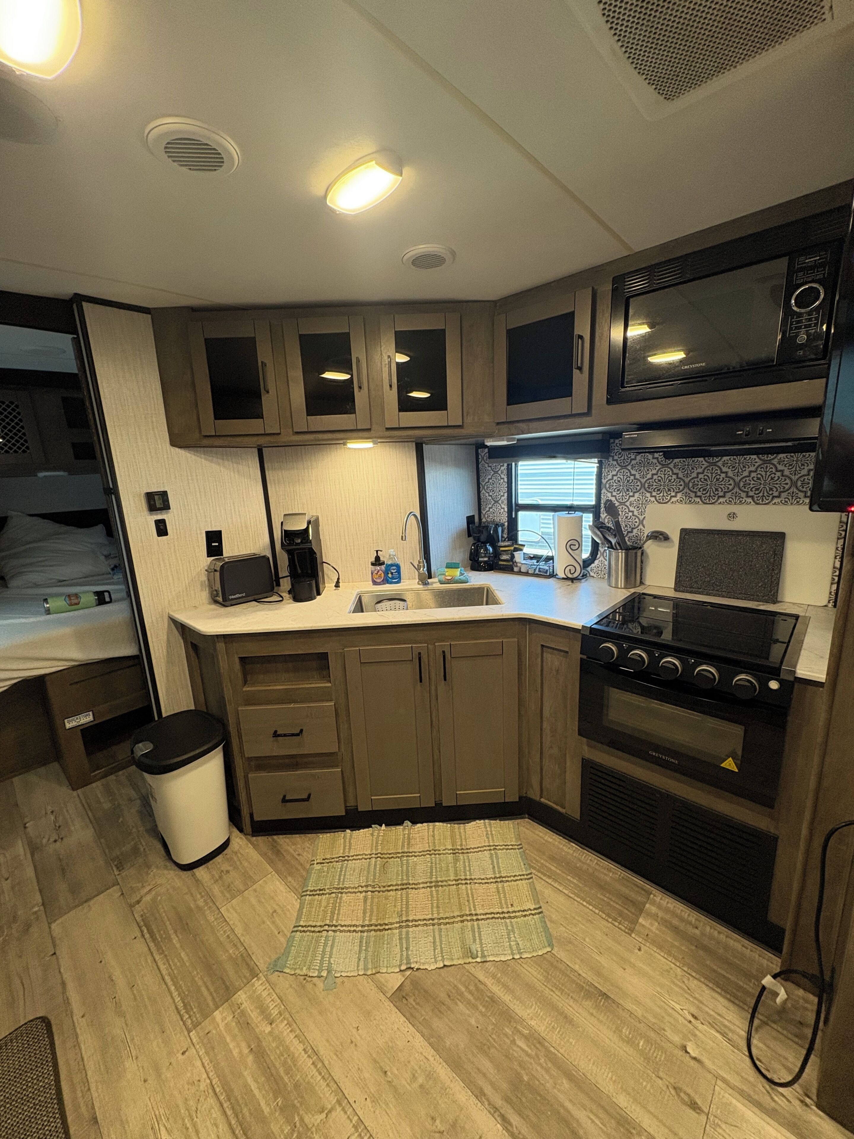 Cocina compacta de autocaravana con estufa, microondas y lavabo, que presenta armarios de madera y una alfombra pequeña.