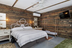 Un dormitorio con una cama, ventilador de techo, aire acondicionado, televisor montado en la pared y una mesita de noche con lámpara.