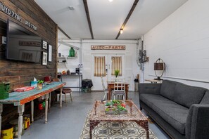 Ein gemütliches Wohnzimmer mit einer Couch, einem Couchtisch mit Pflanzen, einem Fernseher und einer Küchenzeile mit einem Schild, auf dem 'AUTO-PARTS' steht.