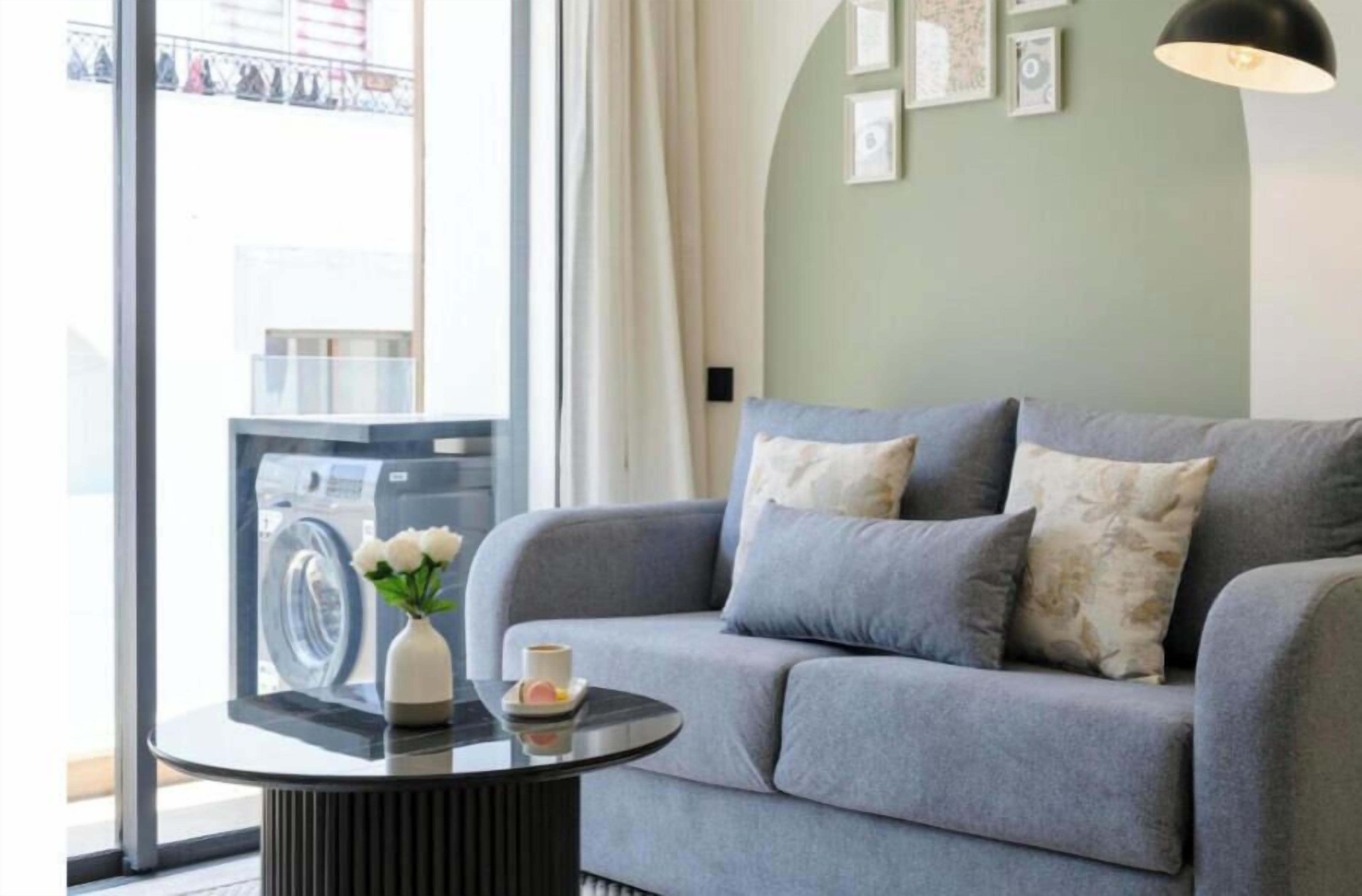 A living room with a grey sofa, a round black table with a vase and a cup, and a large window with a washing machine visible outside.
