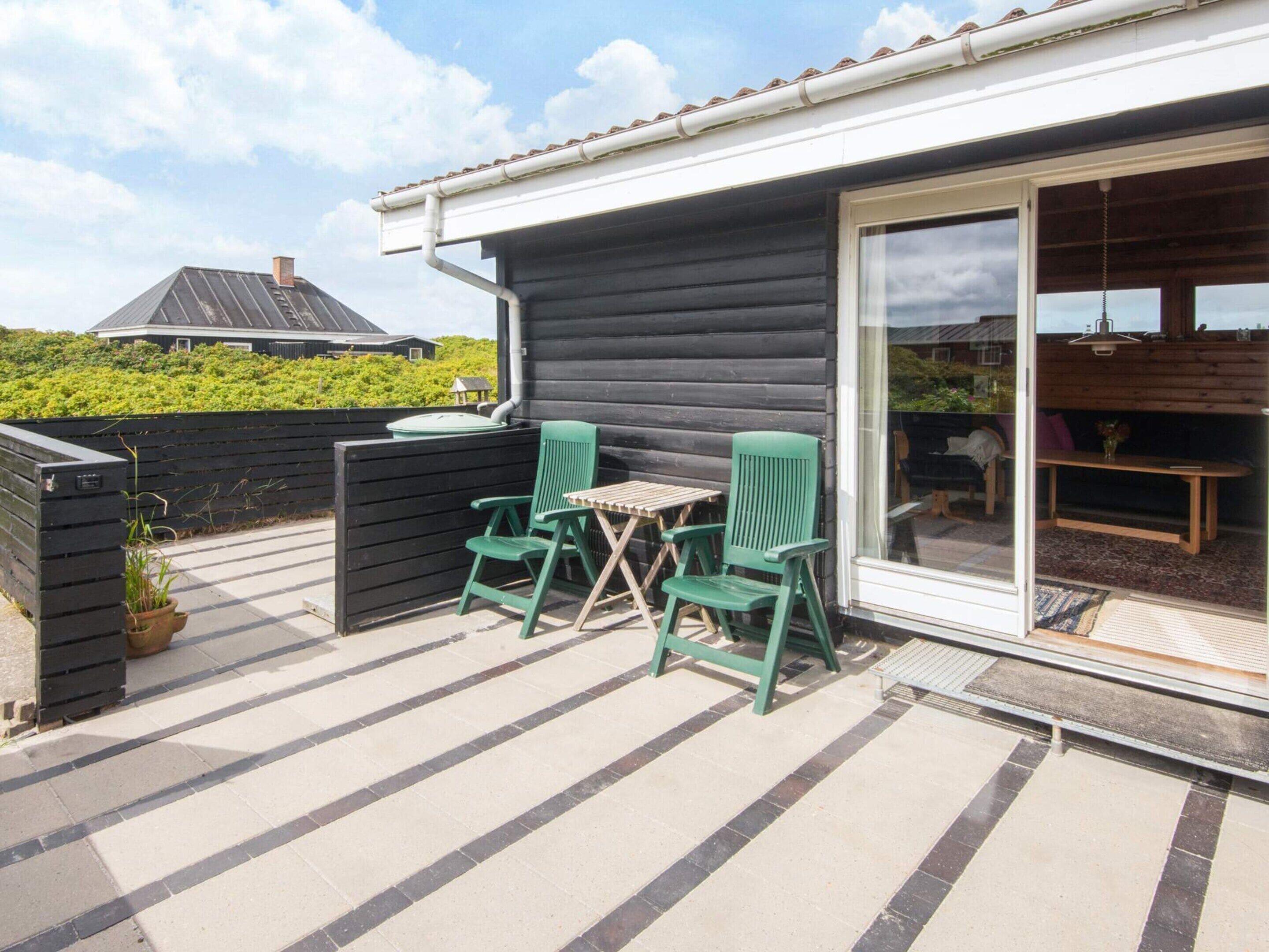 Een terras met twee groene stoelen en een kleine tafel, een glazen schuifdeur naar een binnenruimte, en een uitzicht op een huis op de achtergrond.