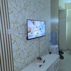 A television mounted on a wall with a geometric pattern, a water dispenser, and a small table with a book. - Cutie Suites TRM | Fast Wi-Fi • Netflix • Parking (Nairobi)