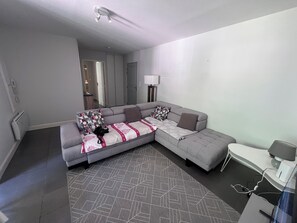 Ein modernes Wohnzimmer mit einem grauen Ecksofa, einem gemusterten Teppich und einem kleinen Beistelltisch mit einer Lampe.