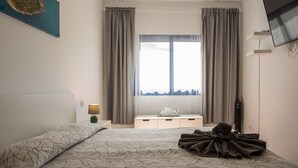 Een slaapkamer met een bed, een nachtkastje met een lamp, een aan de muur bevestigde televisie en een raam met gordijnen.