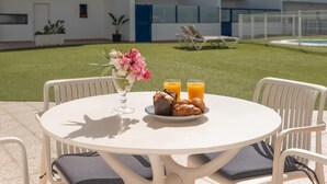 Een ronde buitentafel met witte stoelen, een bloevas, een bord met croissants en gebak, en twee glaasjes sinaasappelsap.