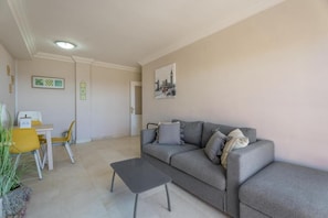 Ein Wohnzimmer mit grauem Sofa, einem kleinen Tisch und einem gelben Sessel. Links steht eine Pflanze und an der Wand hängt ein Bild.