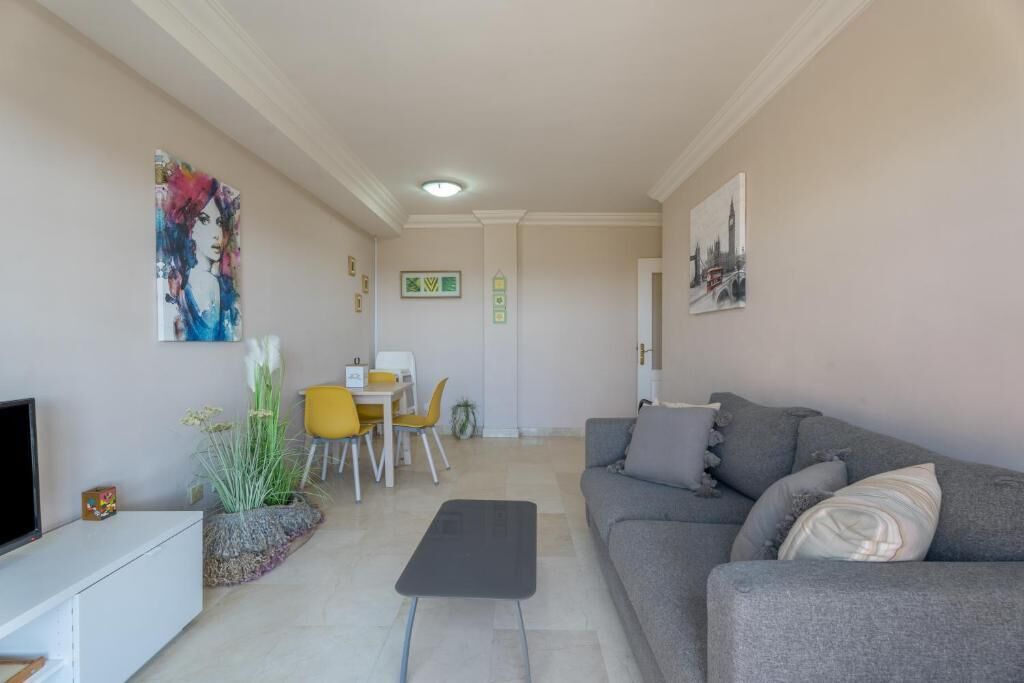 Een moderne woonkamer met een grijze bank, een klein bijzettafeltje en een televisie op een witte kast. Er is een eethoek met een gele stoel en een witte tafel.