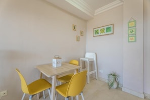 Ein Zimmer mit einem Holztisch, gelben Stühlen, einem weißen Stuhl, einer kleinen Topfpflanze und gerahmten Bildern an der Wand.