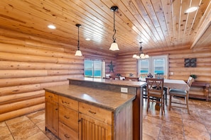 A log cabin interior with a kitchen island, dining area, and large windows offering a view. - Big Bear Cabin | Sleeps 32, Near Marina, Lake Views, AC, Log Cabin! (Garden City)