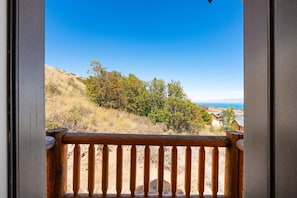 A balcony with a wooden railing overlooking a hillside with trees and a distant view of houses and the ocean. - Big Bear Cabin | Sleeps 32, Near Marina, Lake Views, AC, Log Cabin! (Garden City)