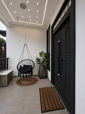 A modern entrance with a black door, a hanging chair, a potted plant, and a textured mat. - 1 + 1 Spa tub. Rooftop Lounge. Airport Pickups/Drop offs (Sakumono)