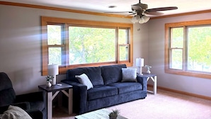 Ein Wohnzimmer mit einem dunkelblauen Sofa, einem Couchtisch und einem Deckenventilator. Fenster bieten Blick auf Bäume.