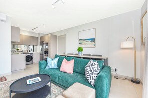 Ein modernes Wohnzimmer mit einer Couch, einem Couchtisch, einem Essbereich mit Tisch und Stühlen, einer Küche mit Geräten und einer Waschmaschine.