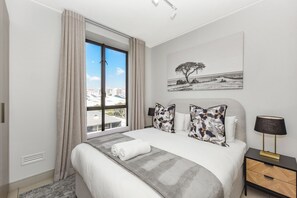 Ein modernes Hotelzimmer mit einem großen Bett, Blick auf die Stadt durch ein Fenster, einem Bild an der Wand und einem Nachttisch mit einer Lampe.