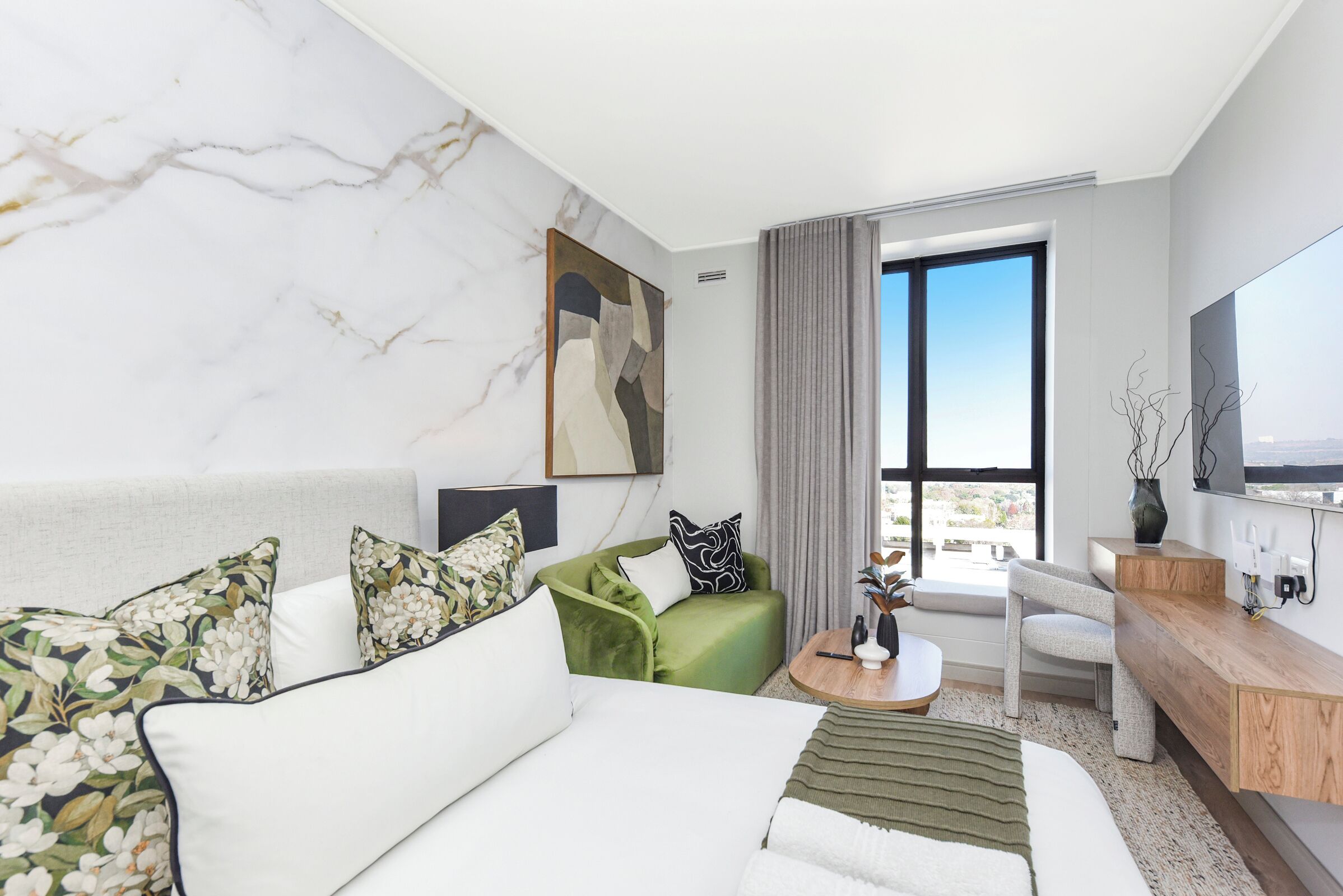 A modern hotel room with a large bed, a green sofa, a small coffee table, a TV, and a large window with a view.