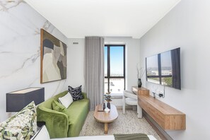 A modern living room with a green sofa, a wooden coffee table, a TV mounted on the wall, and a view of the city through a large window. - One Rosebank: Urban Haven Studio (Johannesburg)