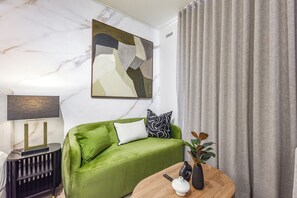 Un canapé vert, un mur en marbre et un tableau, une table ronde en bois avec un vase et une plante, et un rideau gris.
