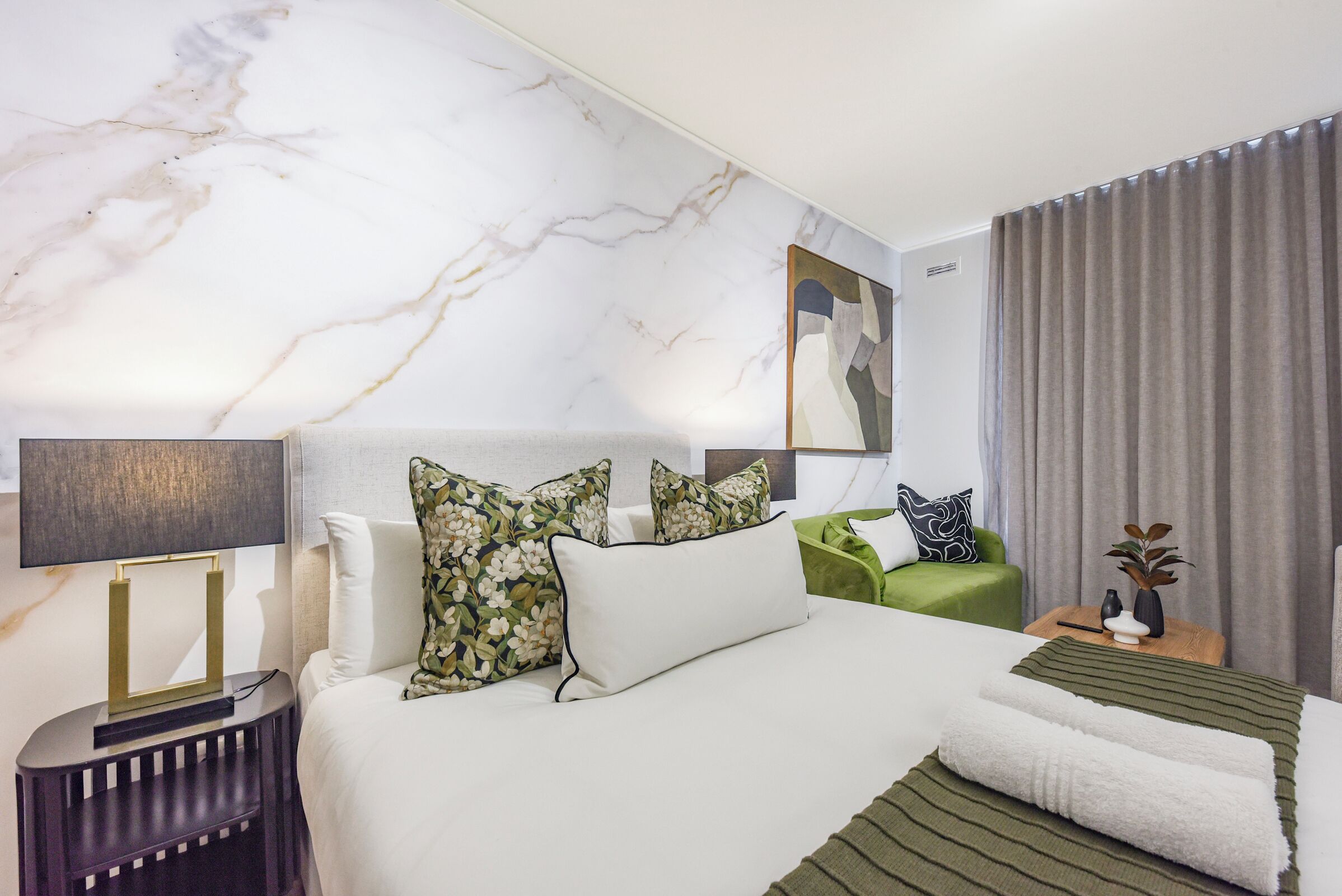 Habitación de hotel con una cama grande, una mesita auxiliar, una silla verde, una lámpara de pared y una cortina.