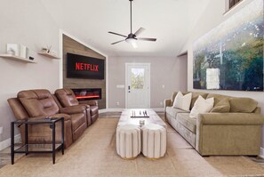 Ein Wohnzimmer mit braunem Ledersofa, einem beigen Couchen, einem Couchtisch, einem an der Wand montierten Fernseher und einem Deckenventilator.