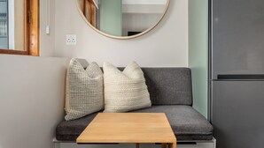 Um espaço compacto para vivenda, com sofá, duas almofadas, uma pequena mesa de madeira, um espelho redondo e um frigorífico.