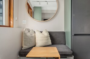 A compact living space with a sofa, two pillows, a small wooden table, a round mirror, and a refrigerator. - Charlie and the Chairman | Contemporary Suite (Cape Town)